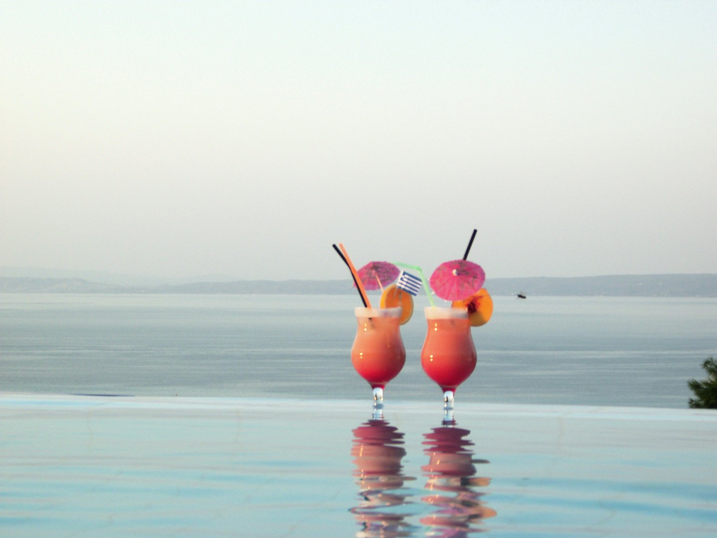 Two tropical cocktails sit by an infinity pool overlooking water.