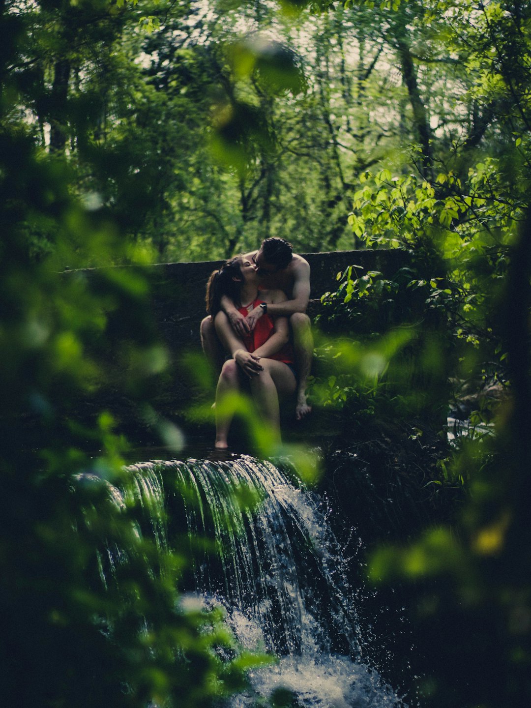 A couple kisses while sitting beside a forest waterfall.